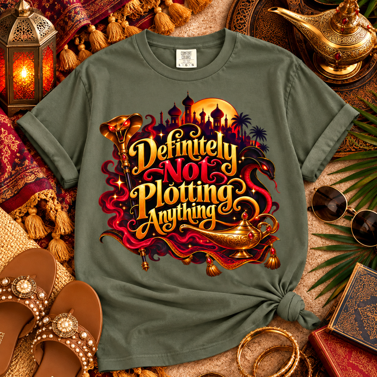 An olive green graphic t-shirt styled in a flat-lay arrangement with Arabian-themed props including a lantern, gold lamp, sunglasses, sandals, and books. The t-shirt features a large graphic with the text 'Definitely Not Plotting Anything' in stylized gold and red lettering, accompanied by a genie lamp and moon design.