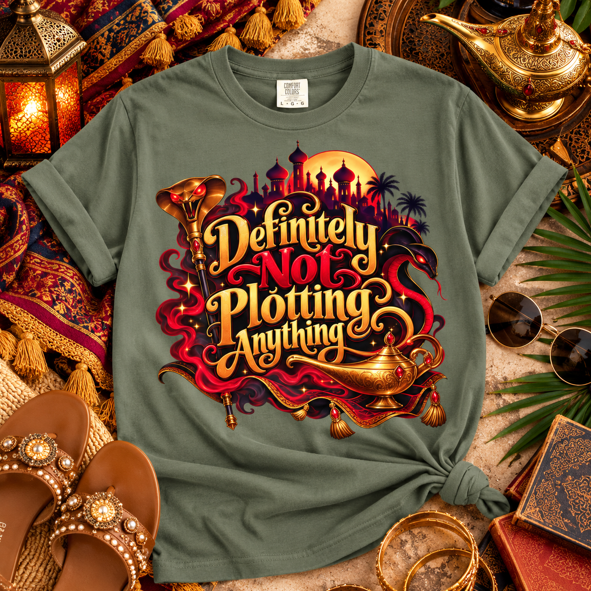 An olive green graphic t-shirt styled in a flat-lay with Arabian-themed props including a gold lamp, sunglasses, sandals, and a lantern. The t-shirt features a large graphic with the text 'Definitely Not Plotting Anything' in stylized gold lettering, accompanied by a genie lamp, a crescent moon, and a desert city skyline.