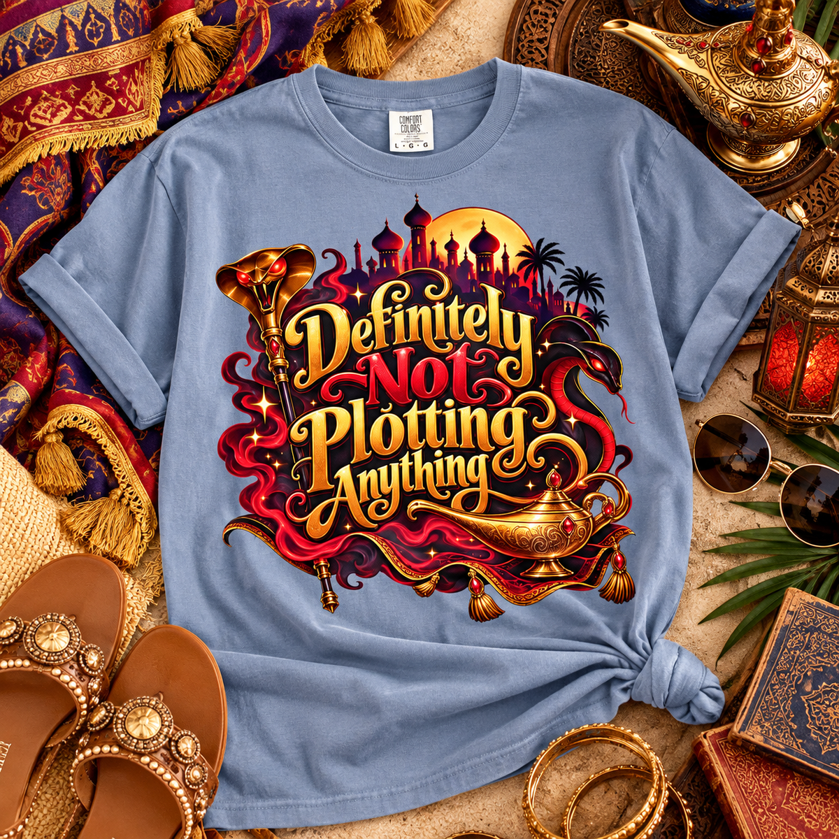 A light blue graphic t-shirt styled in an Arabian-themed flat lay with a gold lamp, sandals, sunglasses, and books. The t-shirt features a colorful graphic with the text 'Definitely Not Plotting Anything' in gold and red script over a stylized genie lamp and moon background.