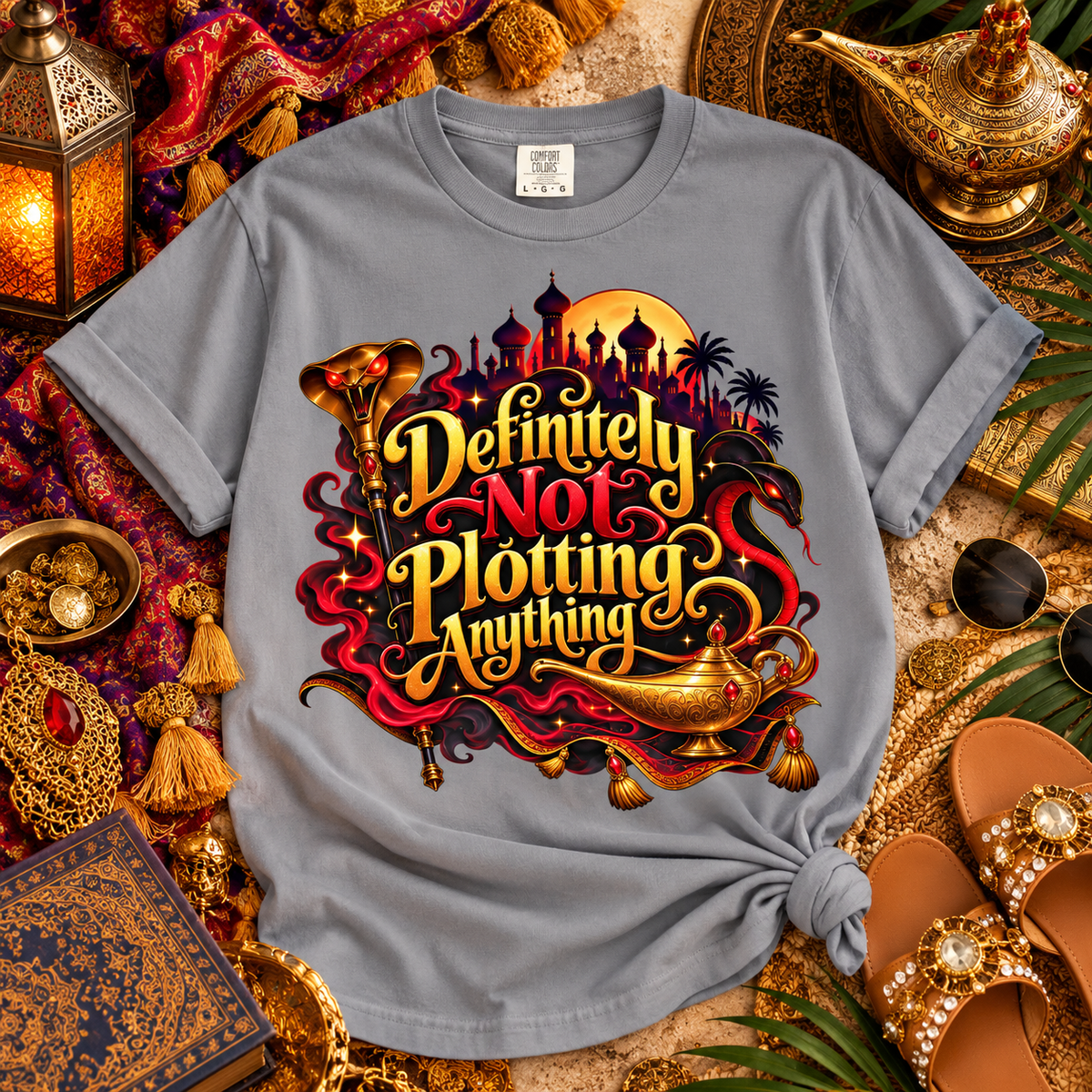 A gray graphic T-shirt laid flat in a styled scene with ornate lanterns, sandals, jewelry, and decorative props. The shirt has a large fantasy-style front print reading “Definitely Not Plotting Anything” in gold and red lettering, with a genie lamp, palm trees, and a moon/sun motif.