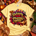 A pale yellow graphic T-shirt laid flat with a styled Arabian-themed scene around it. The shirt features a large front print with the phrase “Definitely Not Plotting Anything” in stylized gold lettering over a red, purple, black, and gold fantasy illustration including a lamp-shaped silhouette and city skyline.