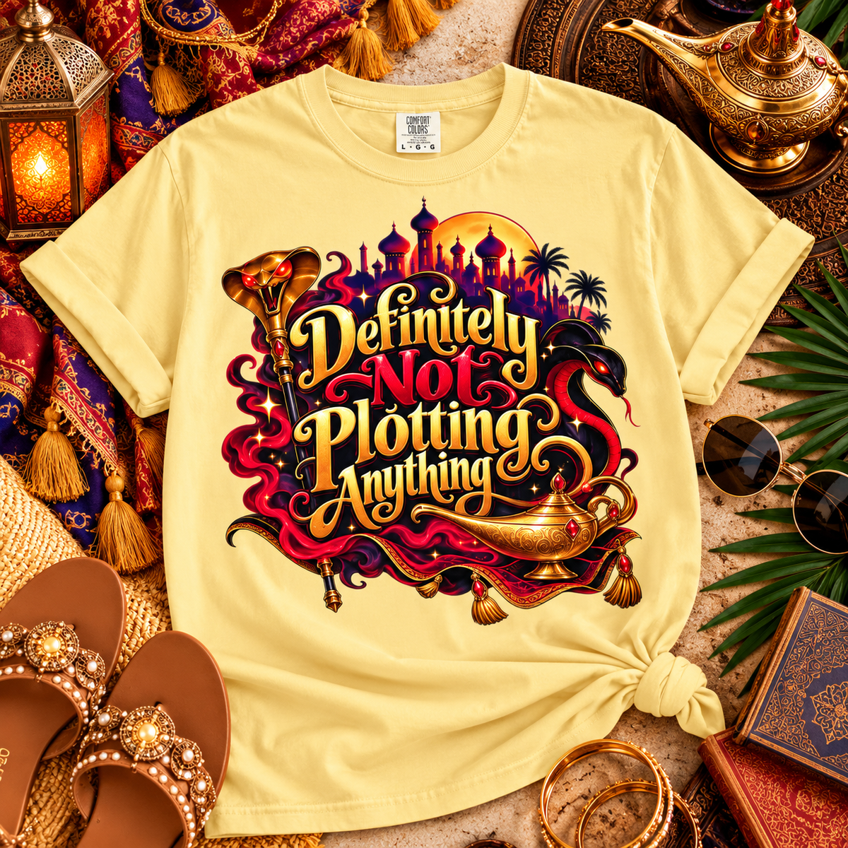 A pale yellow graphic T-shirt laid flat with a styled Arabian-themed scene around it. The shirt features a large front print with the phrase “Definitely Not Plotting Anything” in stylized gold lettering over a red, purple, black, and gold fantasy illustration including a lamp-shaped silhouette and city skyline.