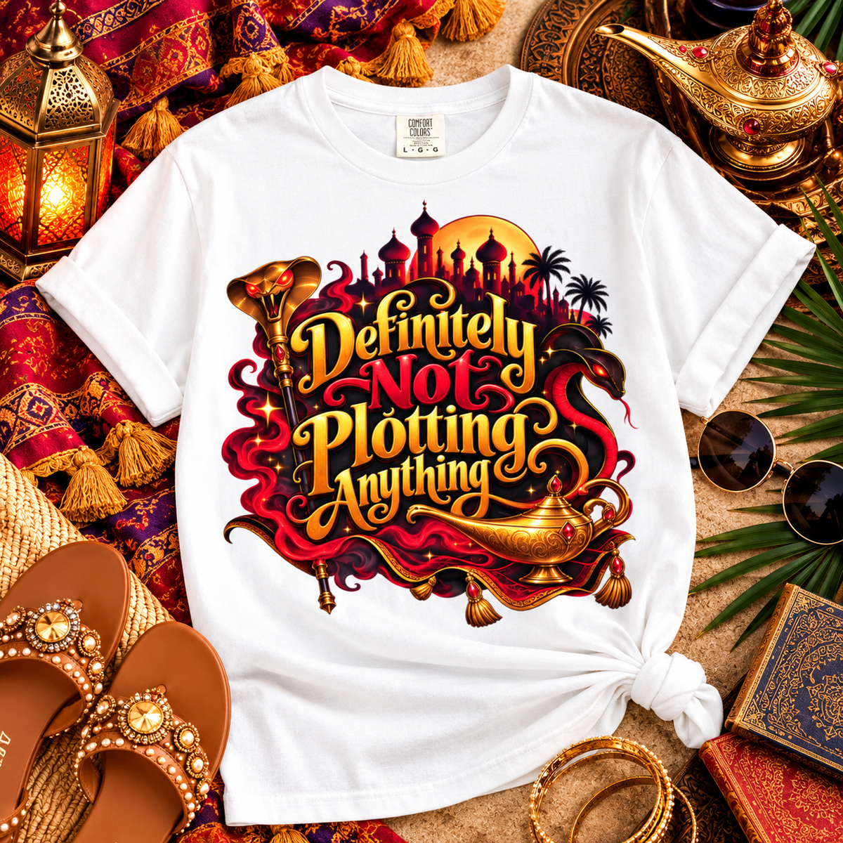 A white graphic T-shirt laid flat on a styled surface with ornate lanterns, sandals, sunglasses, and decorative props. The shirt has a large colorful front print with the phrase “Definitely Not Plotting Anything” in stylized gold lettering over a red, black, and orange fantasy-themed design with a lamp-shaped carafe and skyline silhouette.