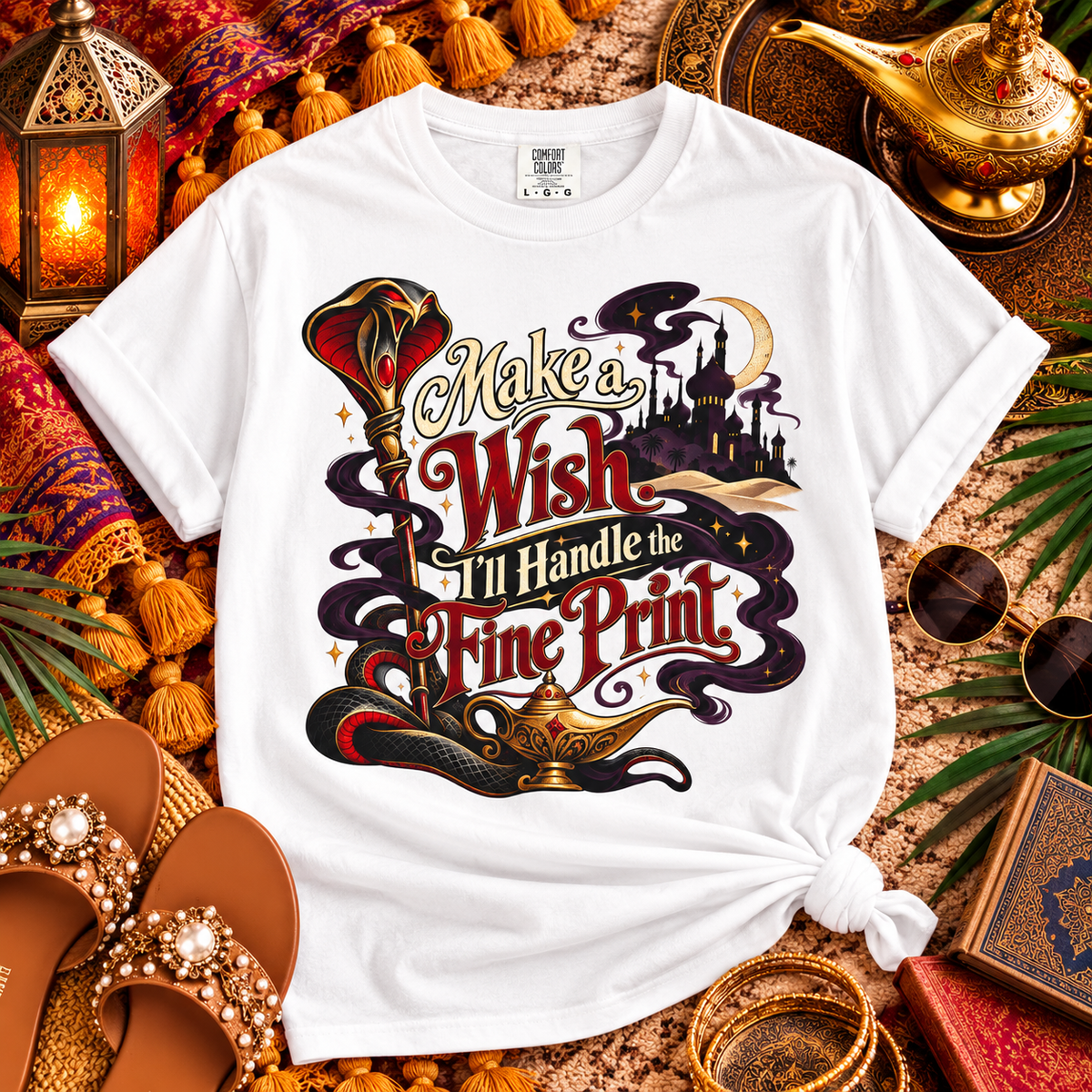 A white graphic t-shirt styled in a flat lay with Aladdin-themed props including a gold lamp, lantern, sunglasses, and sandals. The t-shirt features a colorful graphic with the text 'Make a Wish I'll Handle the Fine Print' and an illustration of a genie lamp and castle.