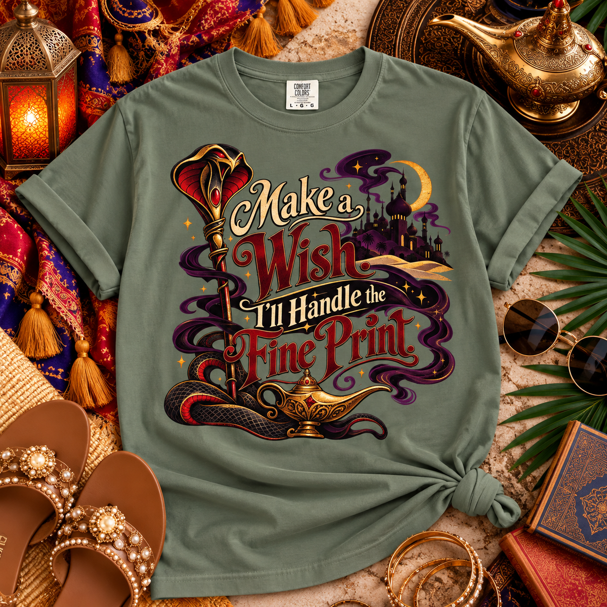 An olive green graphic t-shirt styled in a magical, whimsical flat-lay setting with an Aladdin theme, including a lamp, sunglasses, sandals, and a lantern.