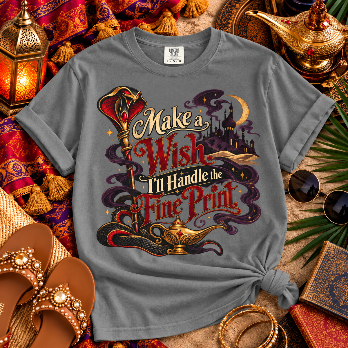 A gray graphic t-shirt styled in a flat-lay arrangement with Aladdin-themed props including a lamp, sandals, sunglasses, and a lantern. The shirt features a colorful graphic with the text 'Make a Wish I'll Handle the Fine Print' and an illustration of a genie lamp.