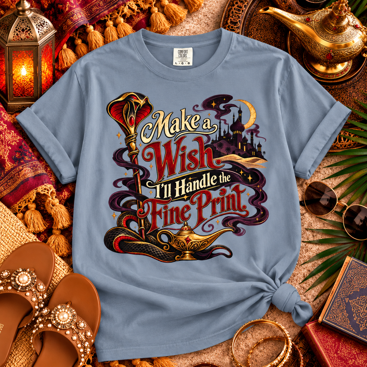 A light blue graphic t-shirt styled in an Arabian-themed flat lay with a keyhole knot at the bottom right. The graphic features a genie lamp, a magic wand, a crescent moon, and the text 'Make a Wish I'll Handle the Fine Print' in stylized red and gold lettering.