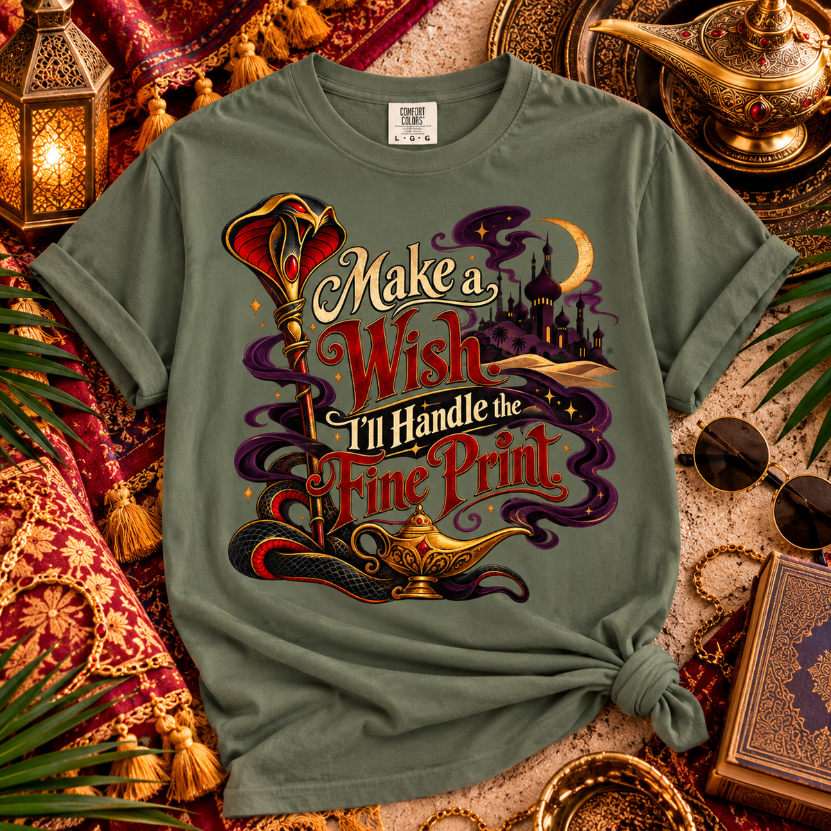 An olive green graphic t-shirt styled in a magical, Arabian-themed flat lay with a lantern, gold teapot, sunglasses, and a book. The t-shirt features a colorful graphic with the text 'Make a Wish I'll Handle the Fine Print' and illustrations of a genie lamp, snake, and castle.