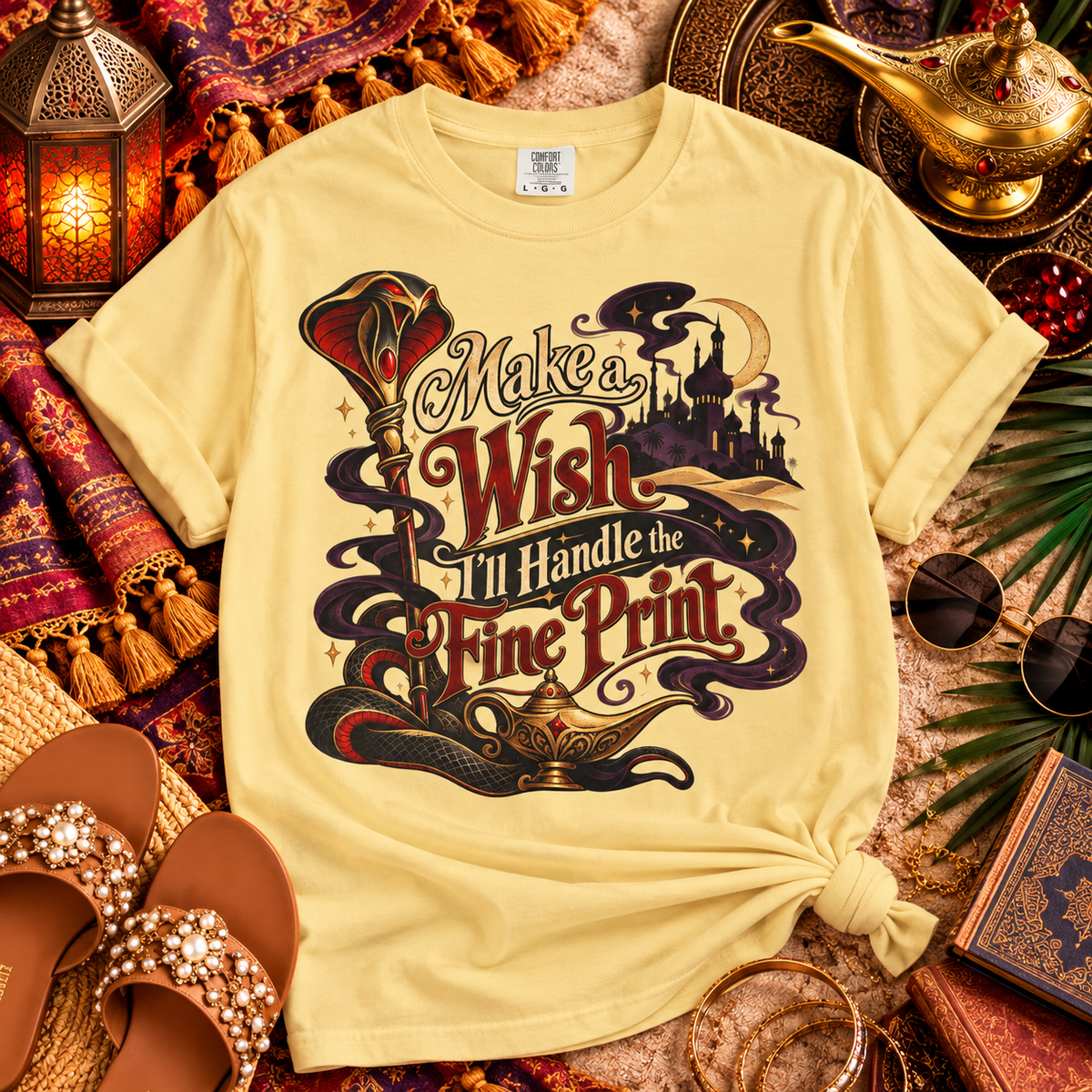 A pale yellow graphic T-shirt laid flat in a styled scene with lanterns, sandals, sunglasses, a lamp, and books. The shirt has a large decorative fantasy-style print reading “Make a Wish I’ll Handle the Fine Print” with a genie lamp, castle, and swirling dark motifs.